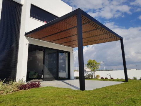 Black aluminum pergola with a wood-look slatted roof on two posts, attached to a modern white home above a paved patio and lawn