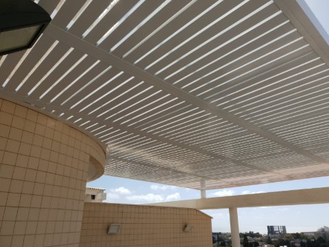 White aluminum louvre pergola projecting from a curved tiled facade, seen from below