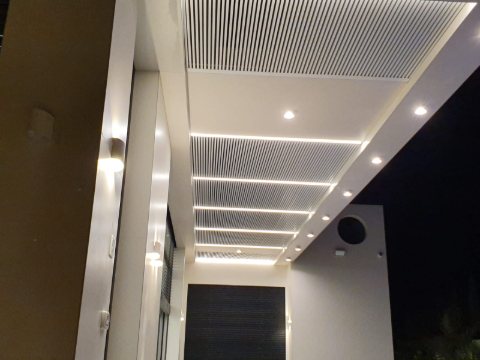 White aluminum louvre pergola with LED strip lights and downlights along a house at night
